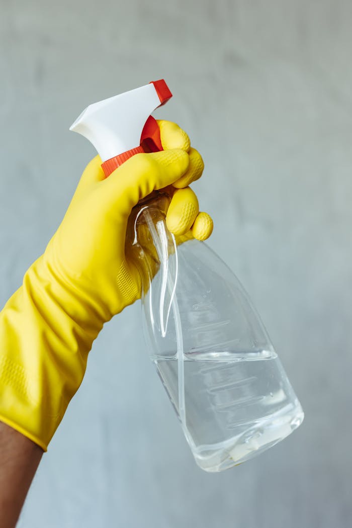 Yellow gloved hand holding a transparent cleaning spray bottle against a neutral background.
